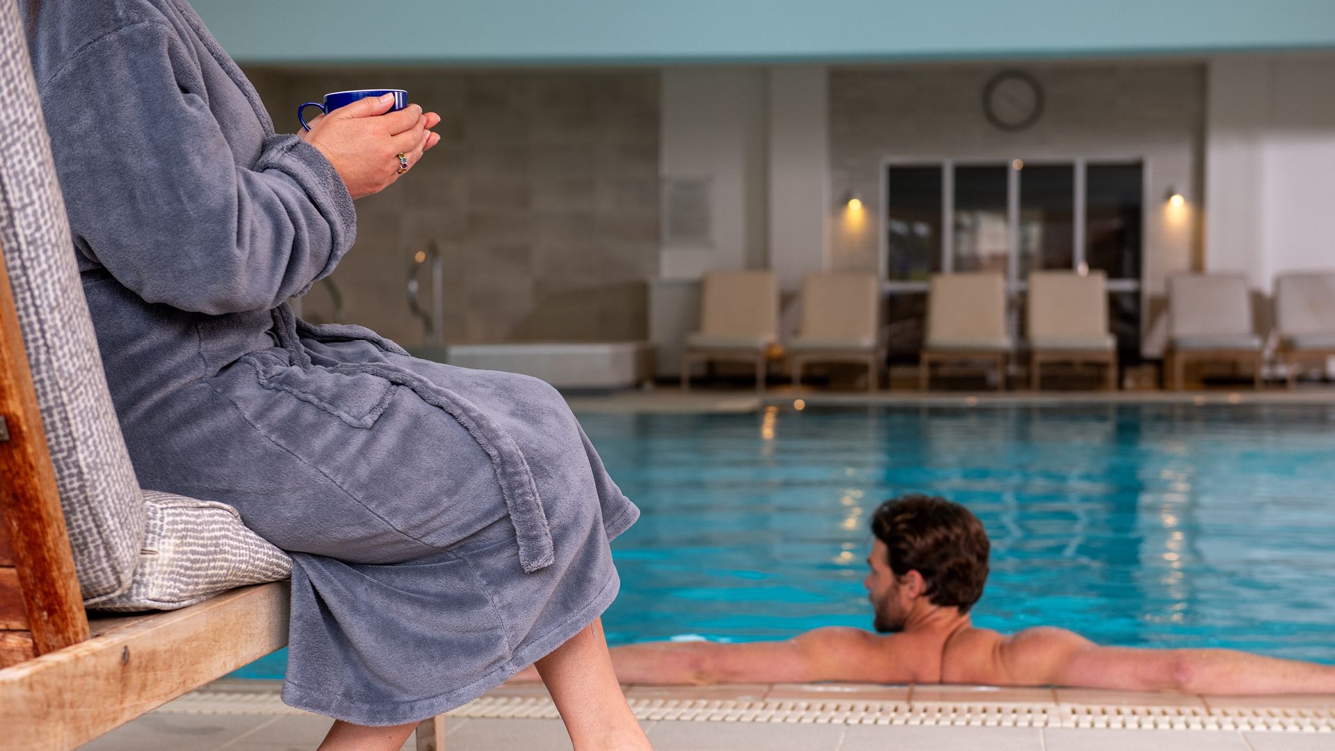 Man in spa pool, woman in spa robe to the left with a hot drink